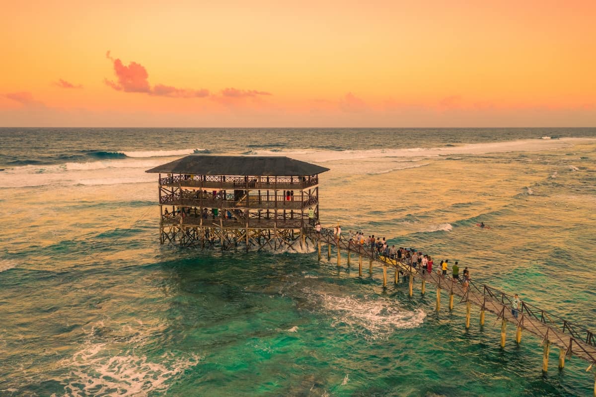 A sunset view of a lone big cottage connected to a bridge and standing on the clear waters with big waves.