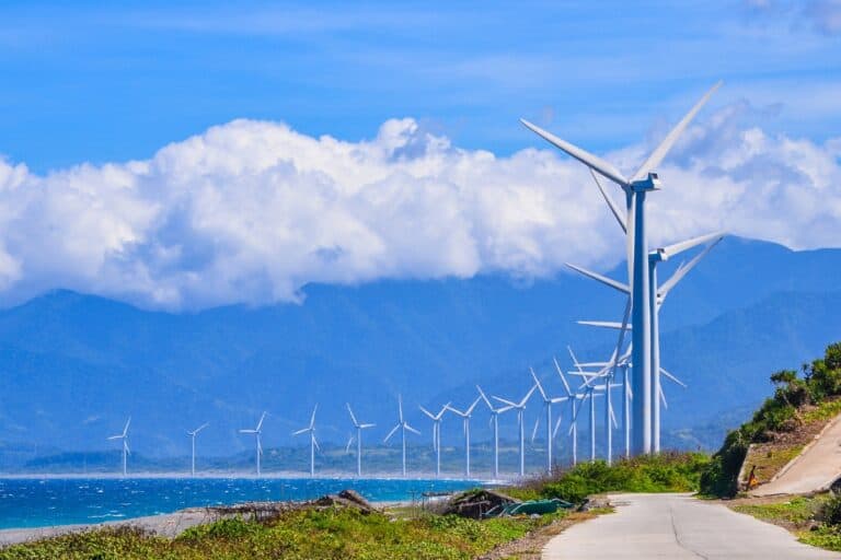 A view of windmills fronting the sea with three propellers each and in the background, mountains and fluffy clouds can be seen.