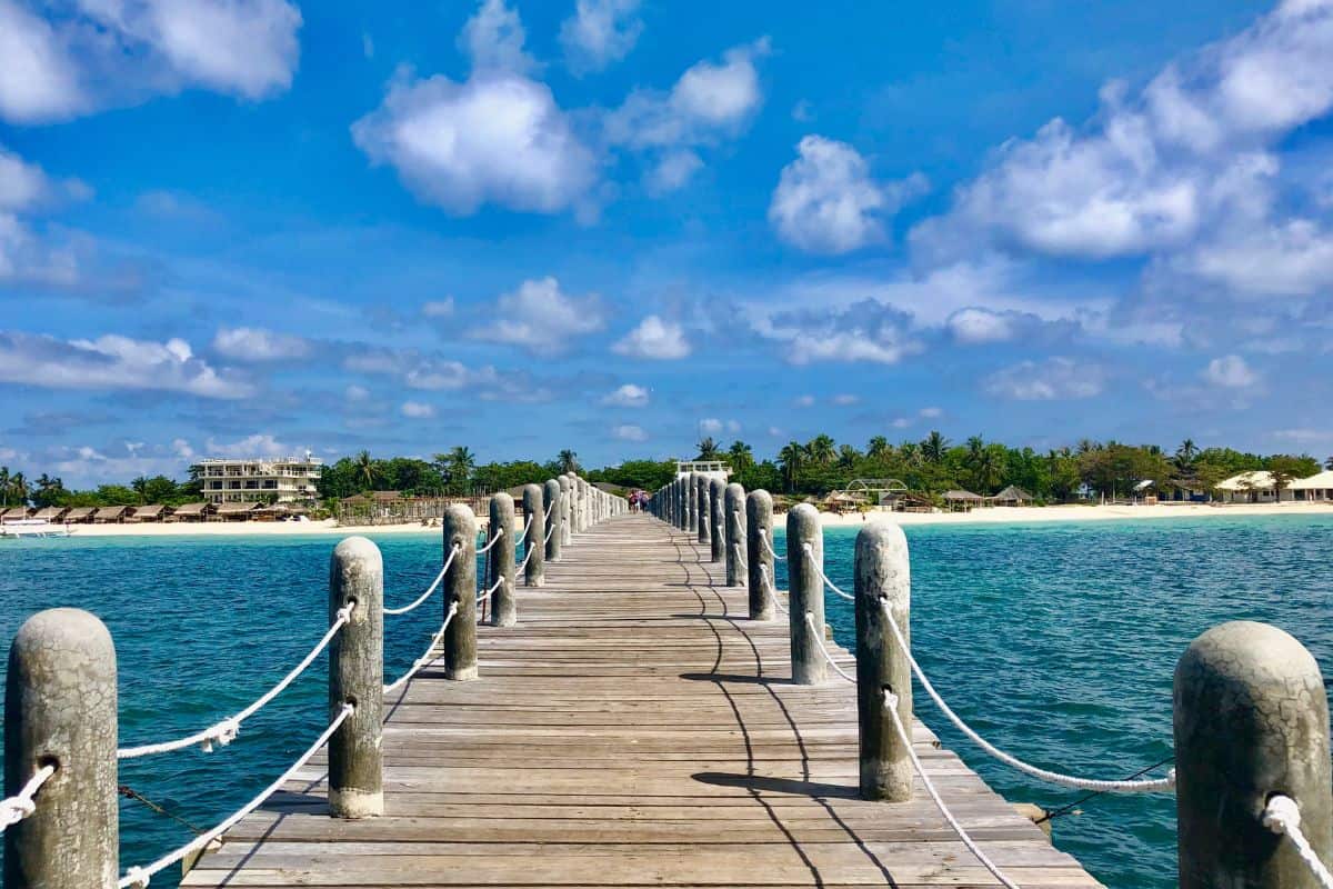 A long walking deck, over a turquoise sea, made of woods, rock, and ropes that ends on a white beach coast with lushes green trees and cottages.
