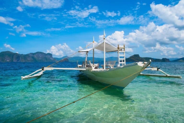 A traditional white outrigger boat floating on clear turquoise water near scenic cliffs, perfect for reaching Hidden Philippine Beaches.