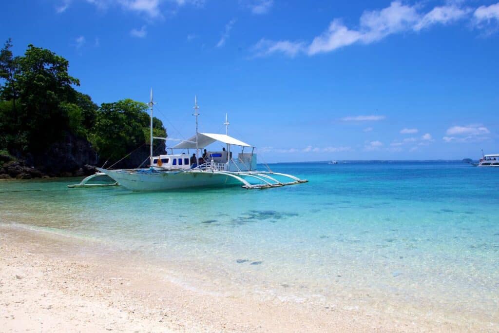 A white outrigger boat anchored near a quiet shore with clear blue water, one of the peaceful Hidden Philippine Beaches.