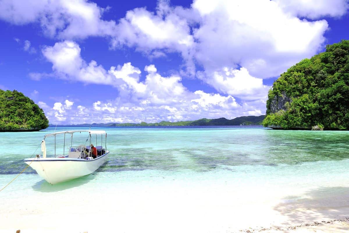 A small white boat anchored on a clear turquoise shore with lush green cliffs in the distance, showcasing the beauty of the Hidden Islands of the Philippines.