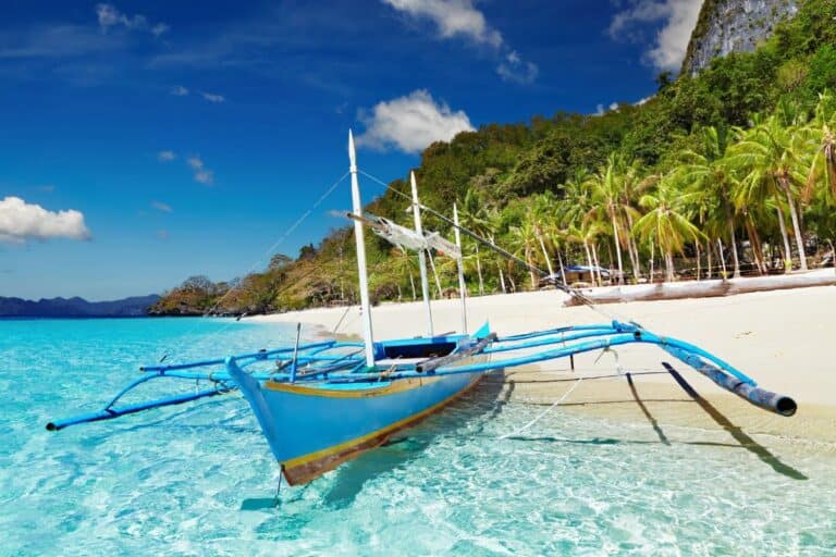 A vibrant blue outrigger boat rests on the clear turquoise shoreline of El Nido, Palawan, with a backdrop of white sand, palm trees, and limestone cliffs—an inviting scene from one of the 5 hidden paradises in the Philippines. Summe rin the philippines one of the Best Season to Visit the Philippines.