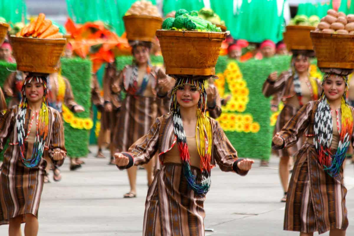 Women in woven tribal-inspired dresses balance baskets of produce on their heads while dancing in a floral parade, highlighting agricultural pride and authentic Filipino culture at the Pagbenga Festival.