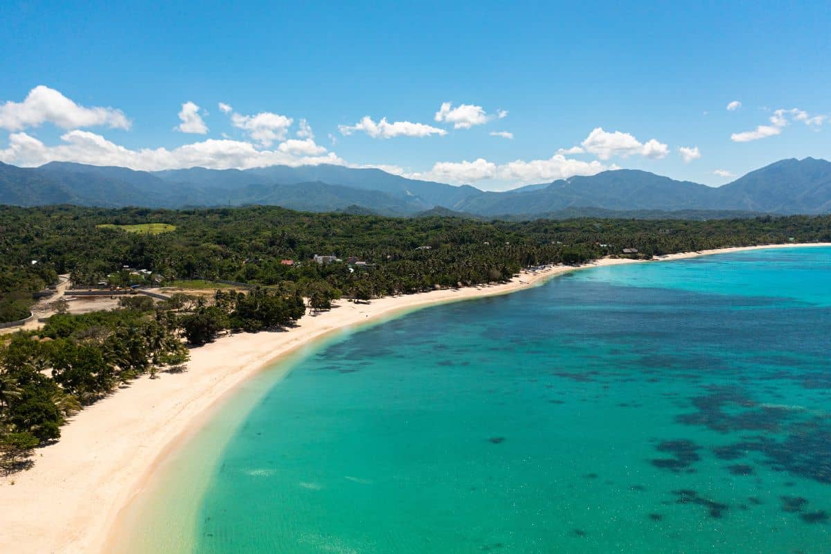 A quiet white sand beach with clear turquoise water and forested mountains in the background, one of the Free Hidden Gems in the Philippines.