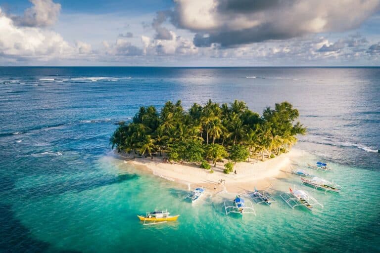 A small tropical island lined with palm trees and surrounded by outrigger boats, one of the Hidden Islands of the Philippines.