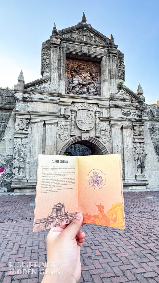 A booklet with stamp being held open in front of the intricately designed gate of Fort Santiago in Intramuros, Manila.