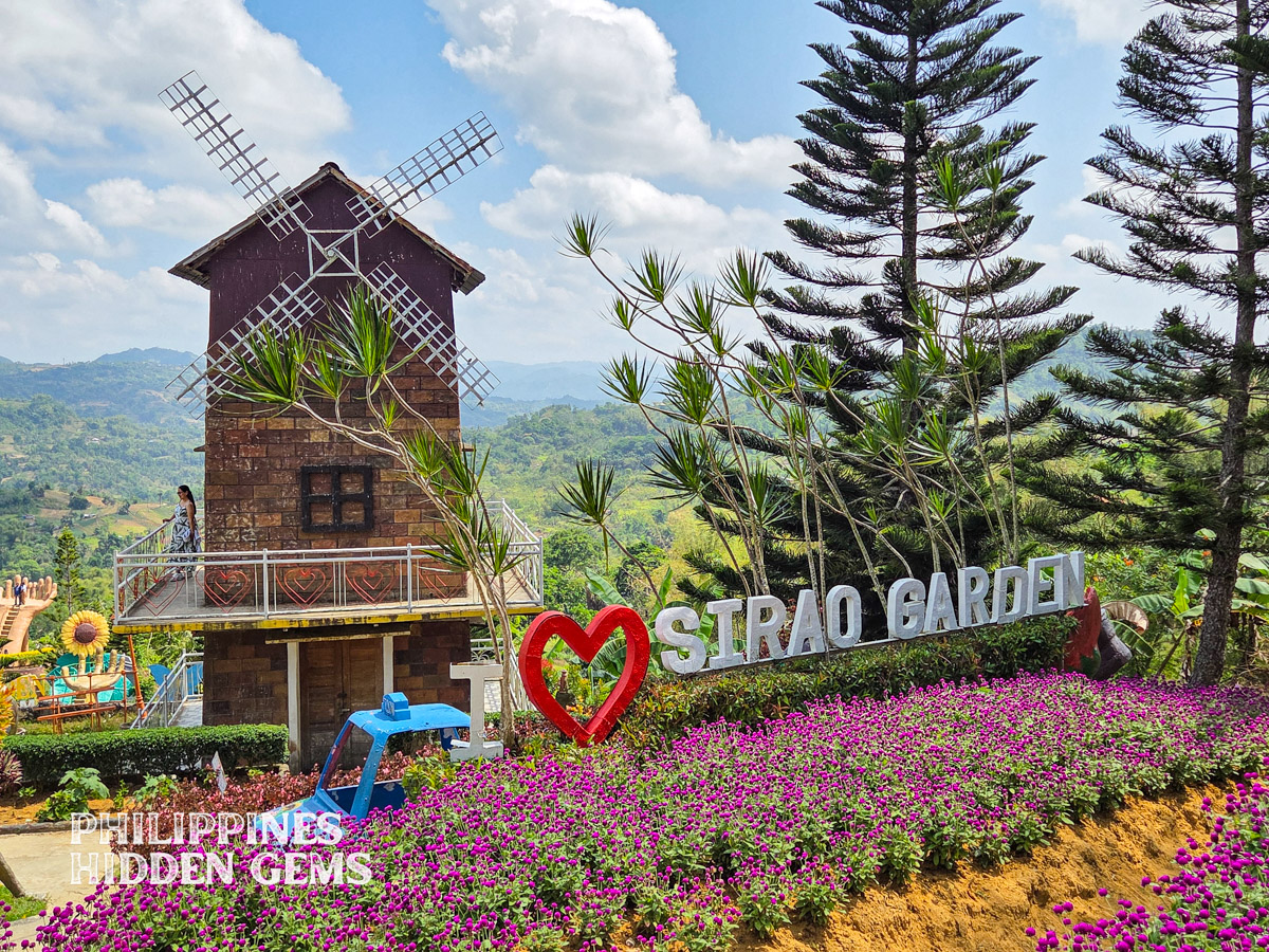 Sirao Garden signage surrounded by colorful flowers with a windmill building and the mountains at the background.