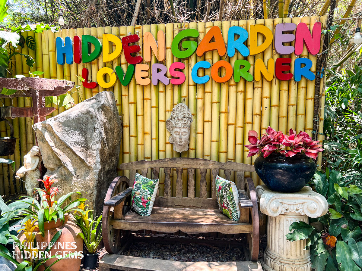 A wooden bench at a garden surrounded by plants, a wall of bamboo wood, and a sign that says "Hidden Garden Lovers Corner".